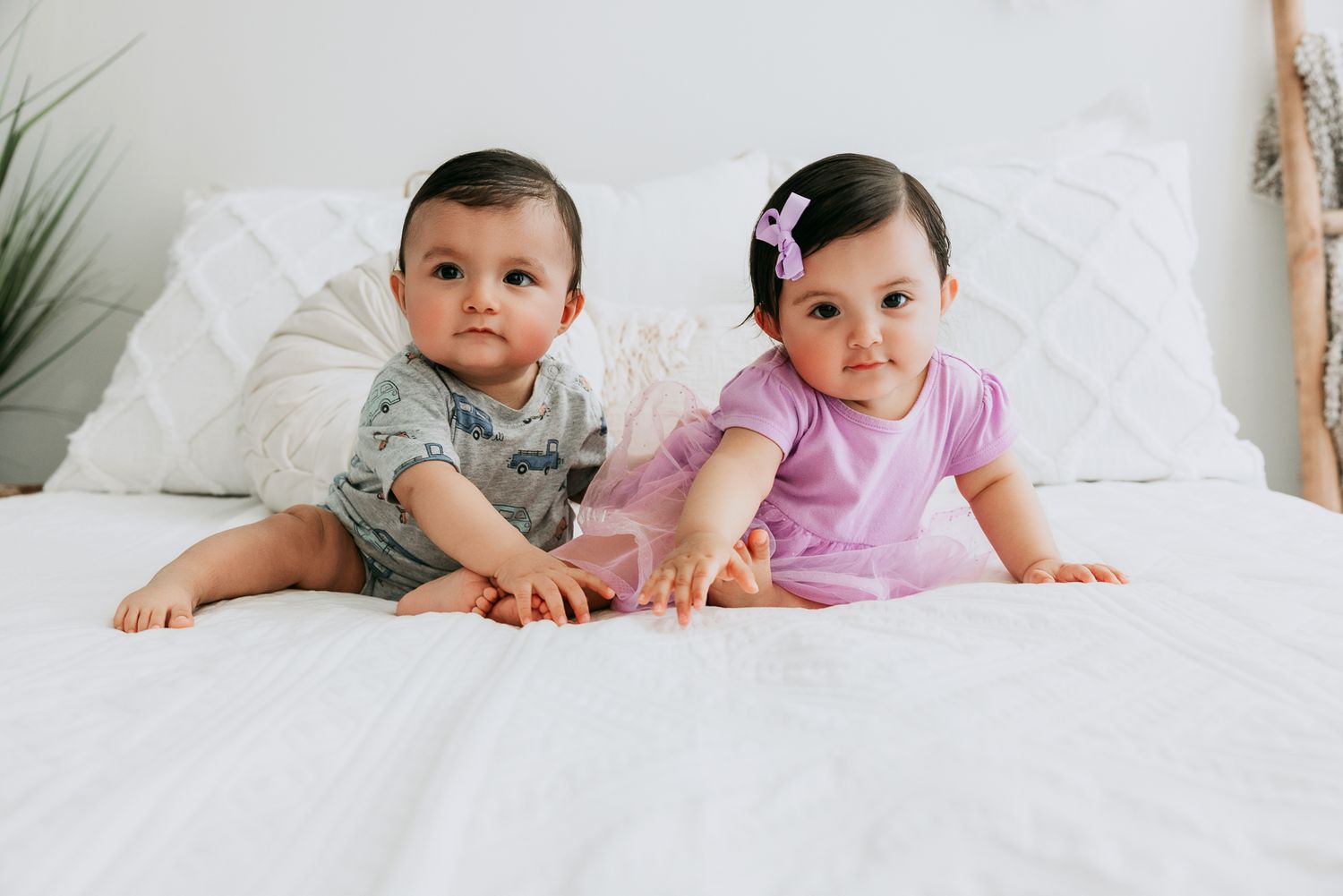Two babies in casual outfits lying together on a white bed, boy in gray and girl in purple.