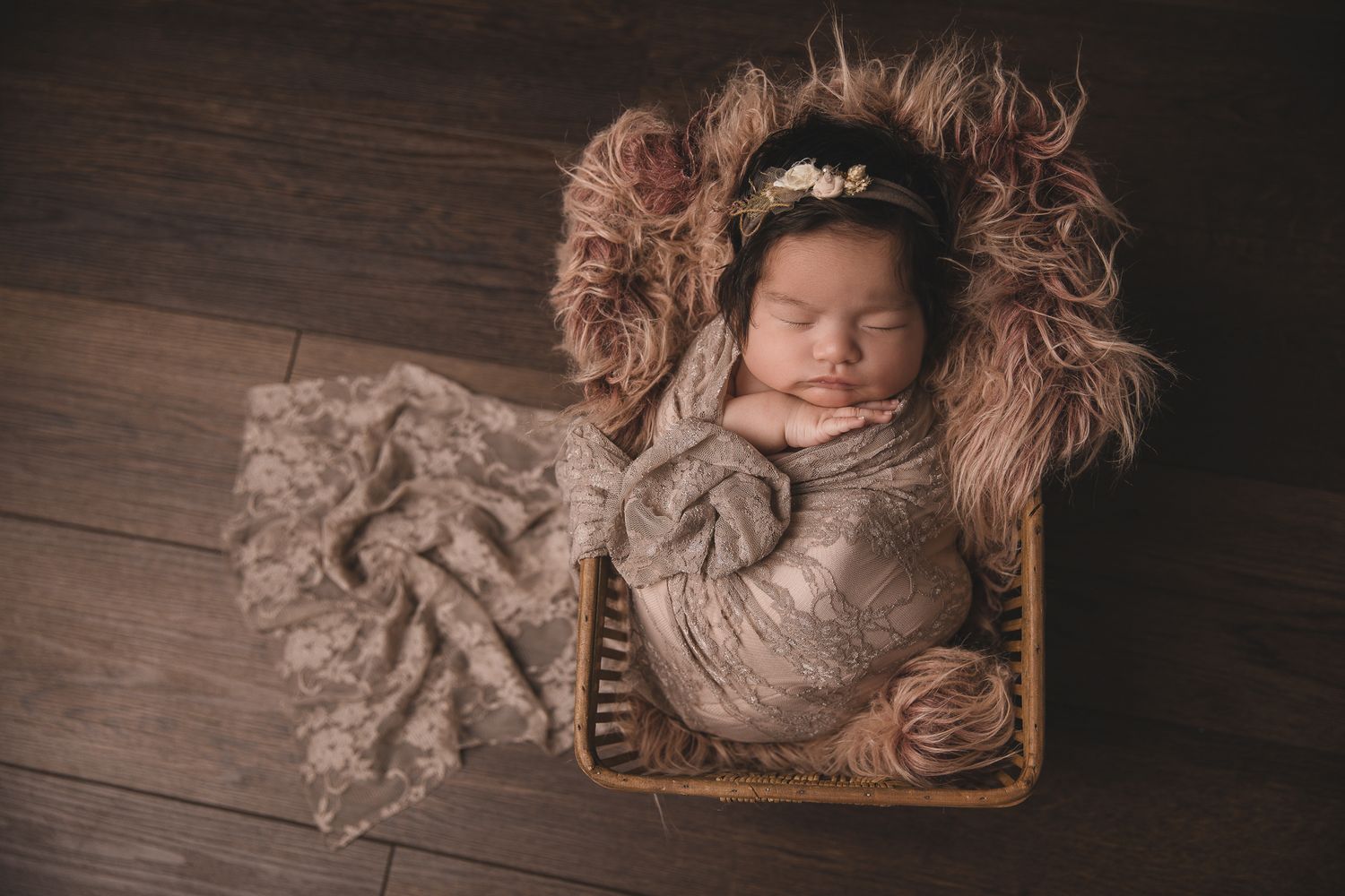 Sleeping newborn baby girl wrapped in taupe lace poses in a wicker basket with fluffy fur in Carlsbad near Bonsall