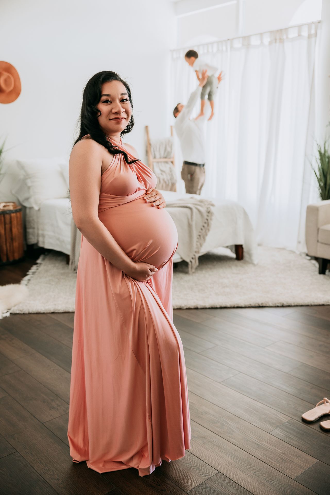 Smiling pregnant woman in pink maxi dress holds baby bump while family plays in bright bedroom background.
