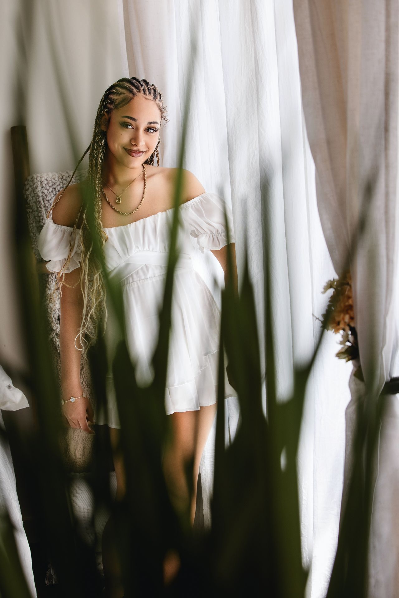 Young woman in white off-shoulder dress with long braids smiles standing between sheer white curtains. Senior photography session in studio