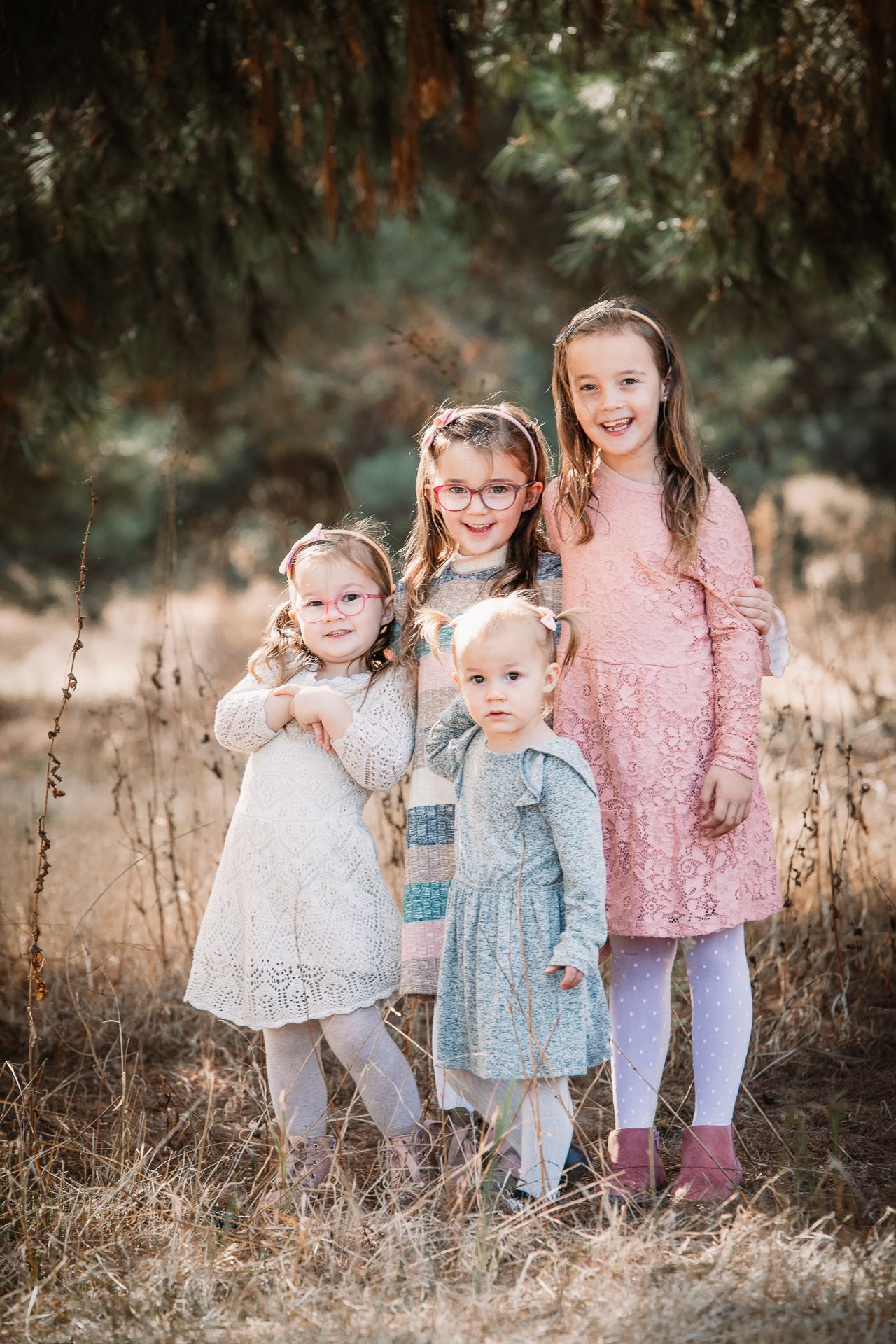 Four young girls in dresses pose together in a golden field surrounded by evergreen trees.