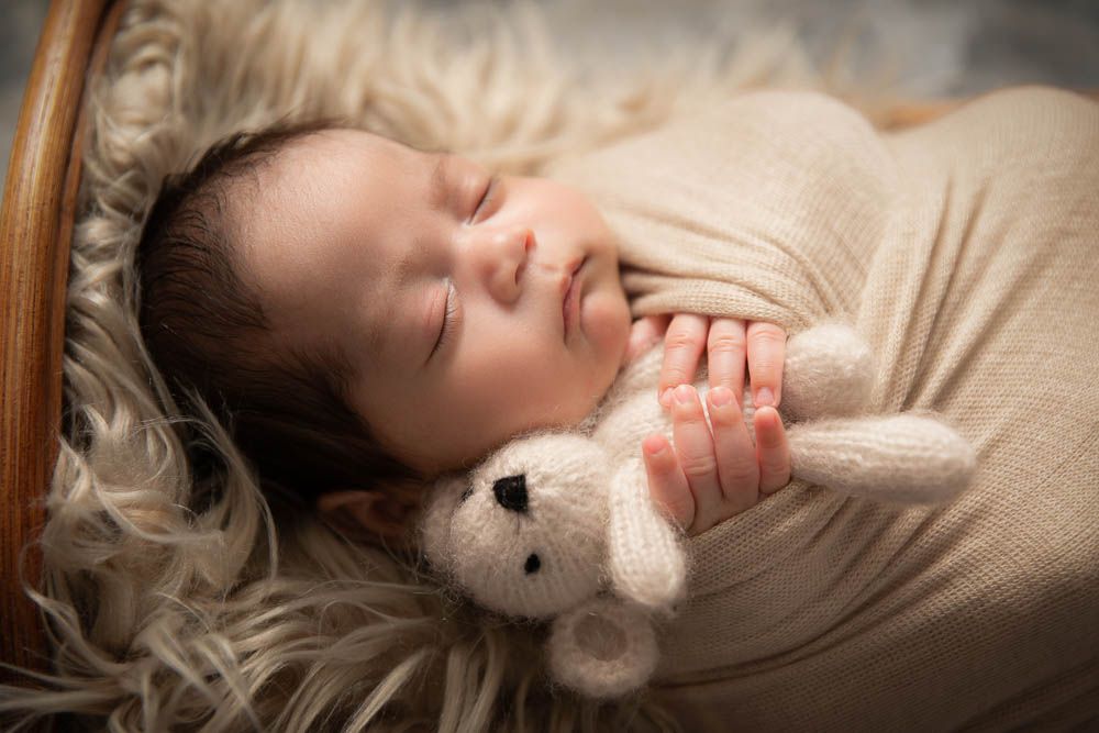 Sleeping newborn baby wrapped in cream swaddle clutching a small white knitted teddy bear in a cozy basket.