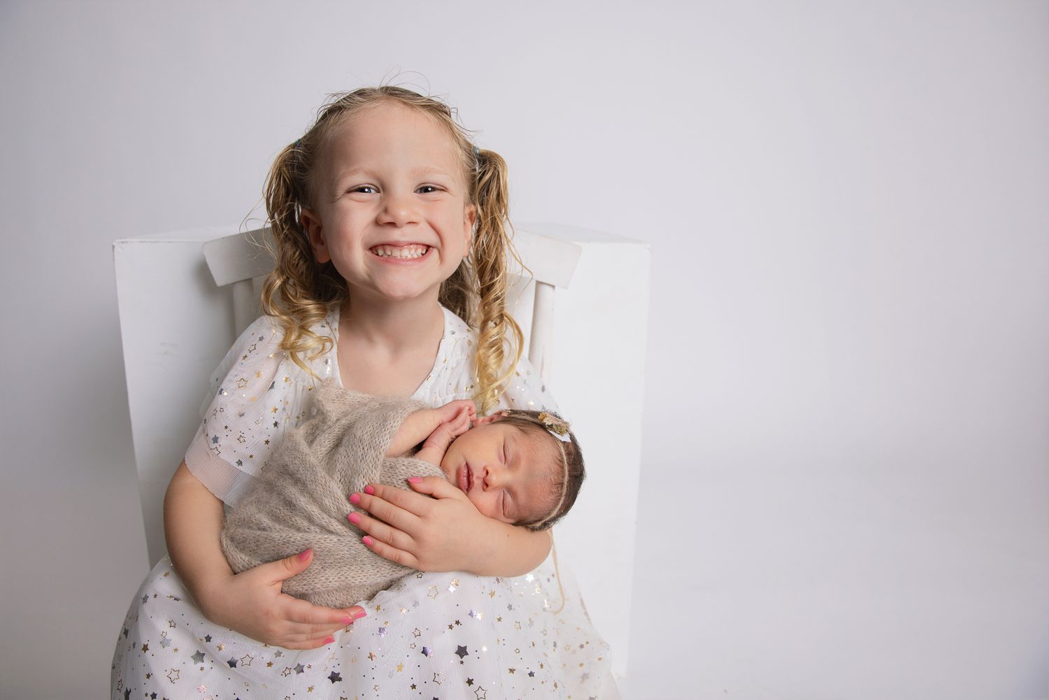 Smiling young girl with curly blonde hair holds sleeping newborn baby wrapped in beige swaddle on white chair.