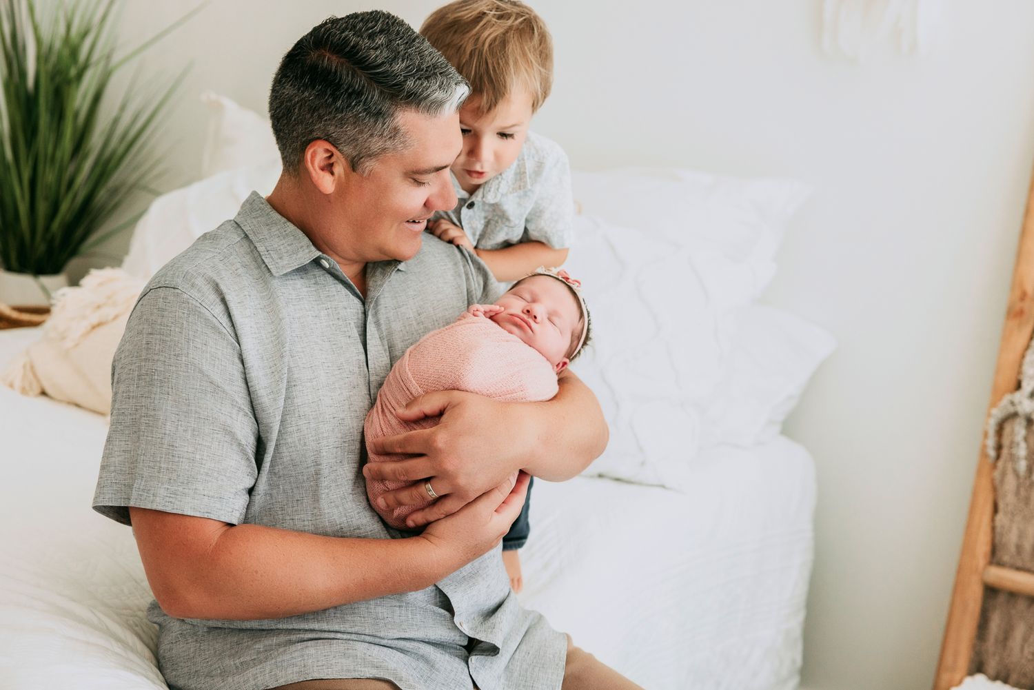 Father holding newborn baby while young toddler son looks on, sitting on white bed in bright room.