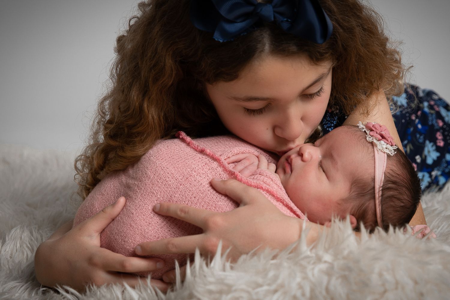 Older sister gently kissing newborn baby wrapped in pink on a white fluffy blanket.
