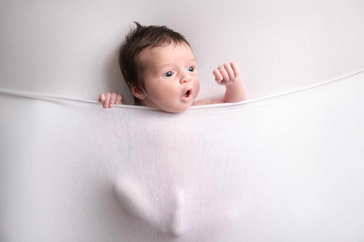 Adorable baby peeking over a white fabric hammock with wide eyes and open mouth on white background.