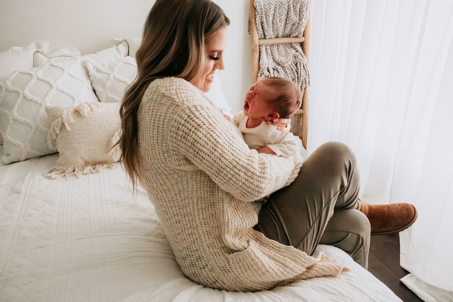 A mother in a cozy knit sweater sits on a bed cradling her newborn baby in a soft, neutral-toned bedroom.