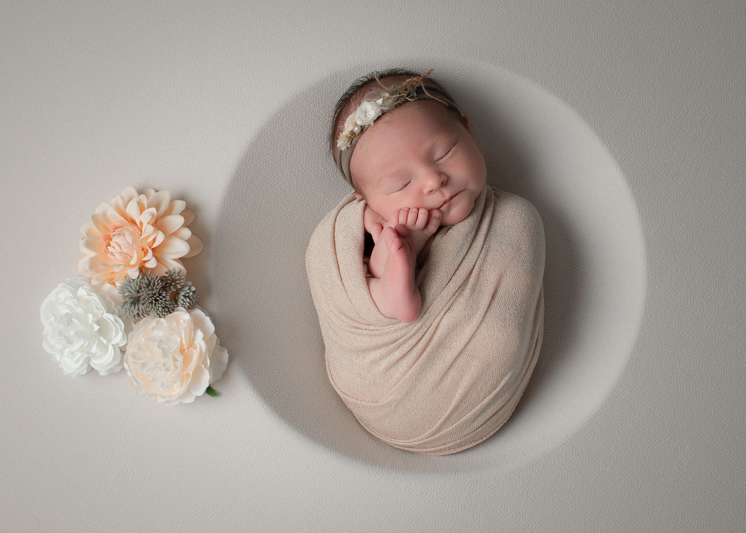 Sleeping newborn baby swaddled in beige wrap with floral headband, posed in white bowl with peach and white flowers.