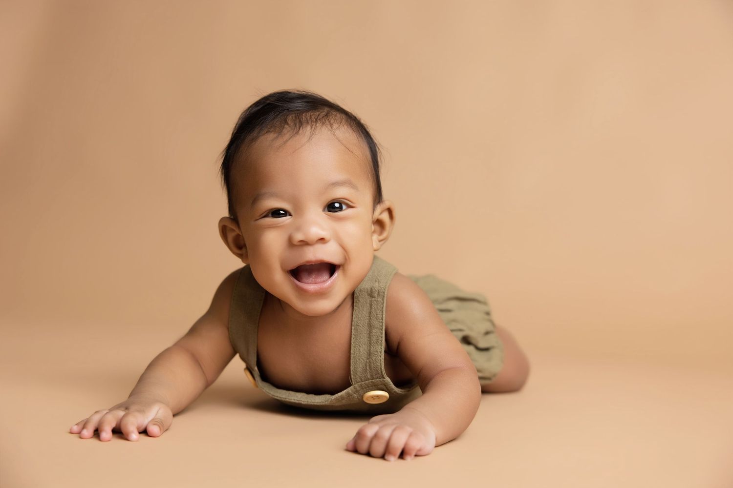 Smiling baby in green overalls doing tummy time on a beige background in a professional studio portrait.