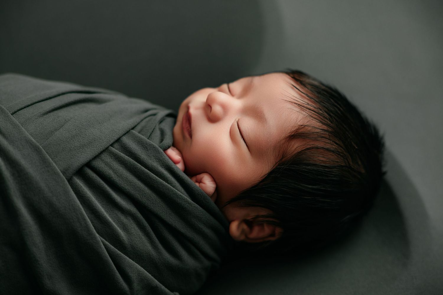 Peaceful newborn baby swaddled in a dark green wrap, sleeping soundly with dark hair visible.
