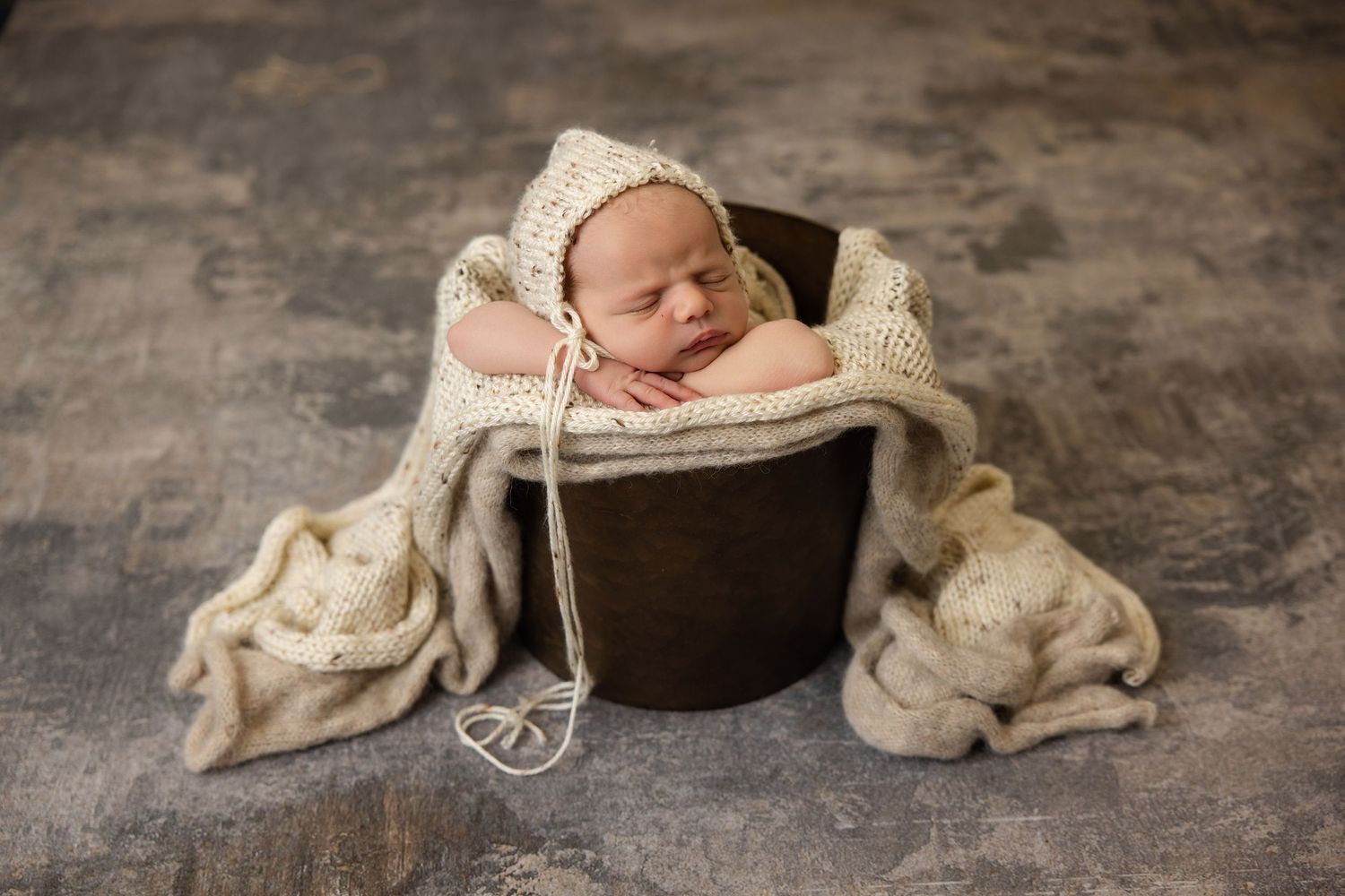 Sleeping newborn baby in cream knit bonnet curled inside a rustic wooden bucket with soft beige blankets.
