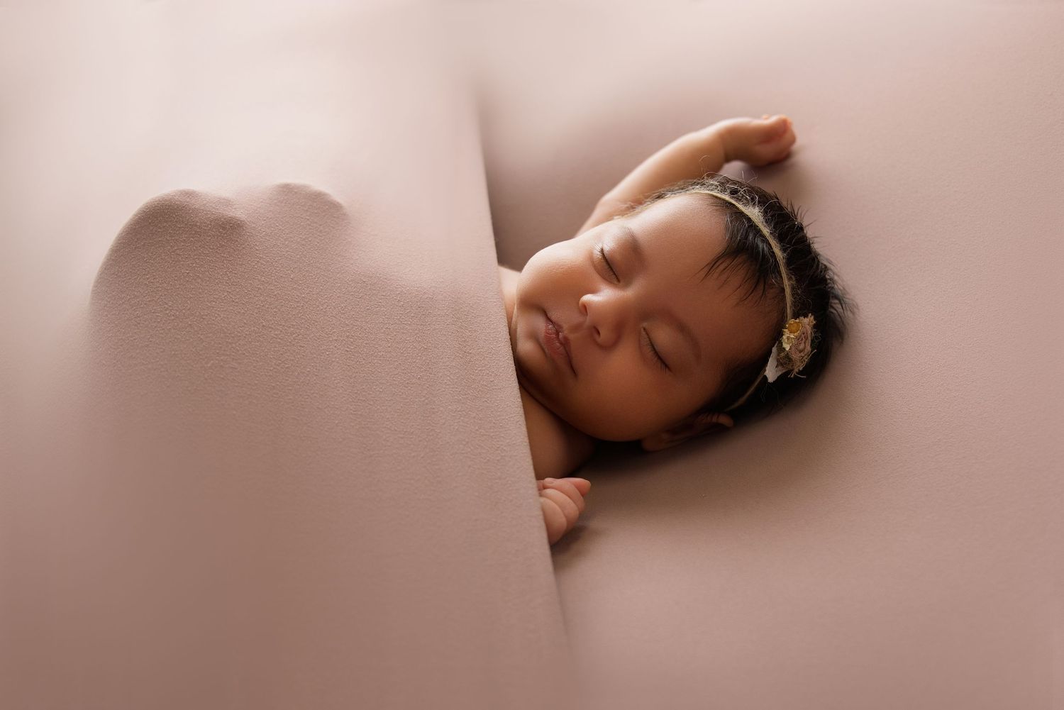 Sleeping newborn baby with a headband nestled between soft pink cushions in a studio photo.