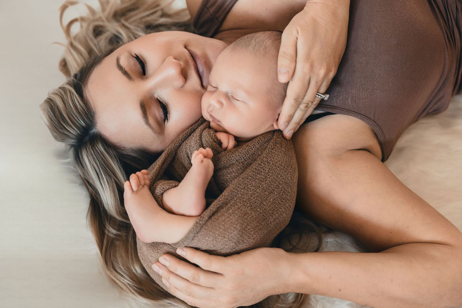 Mother lying down cuddling her sleeping newborn baby wrapped in a brown swaddle cloth.