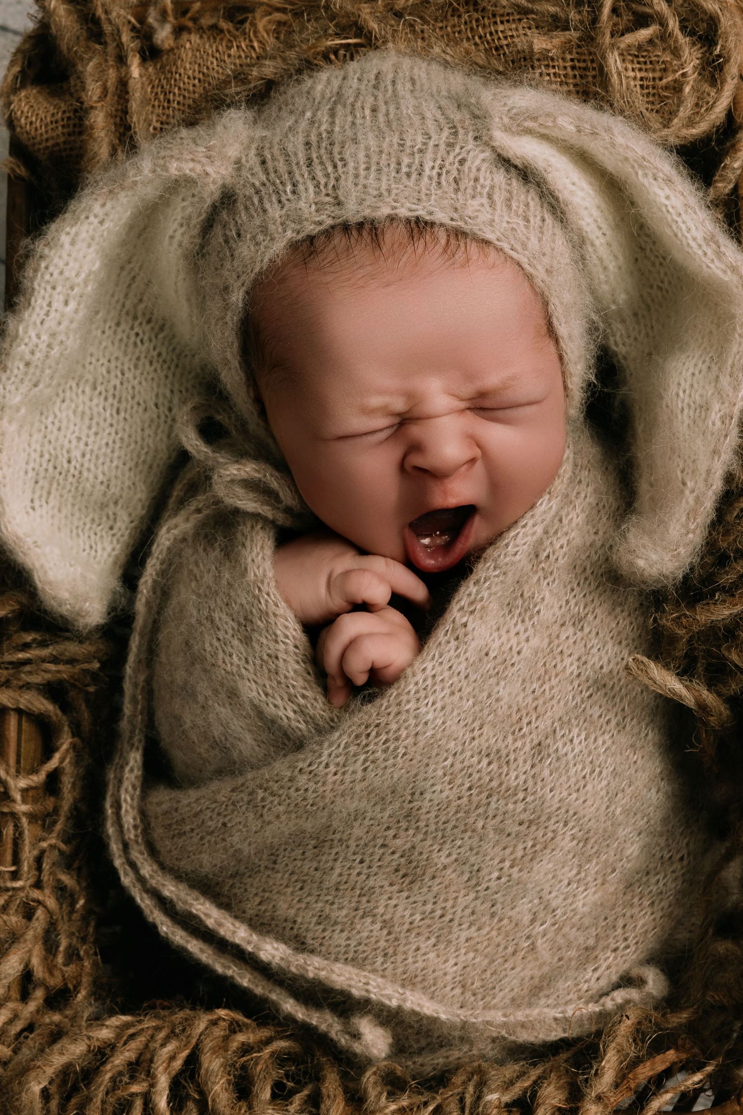 Newborn baby yawning wrapped in a cozy knit blanket wearing a bunny ear hat, nestled in a basket.