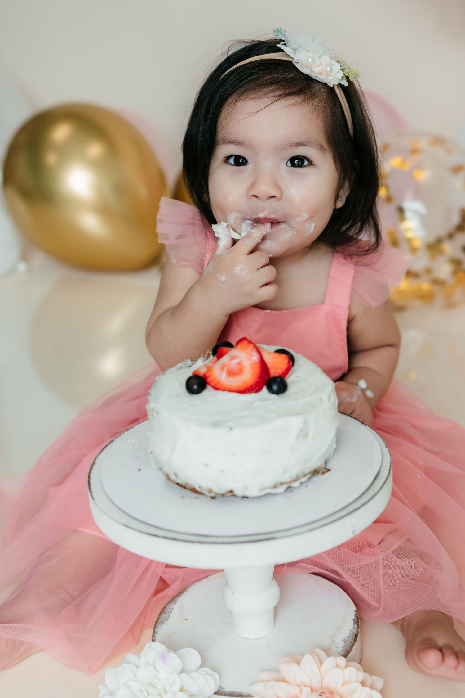 Toddler girl in pink tutu dress eating birthday cake at cake smash photo session with gold balloons.