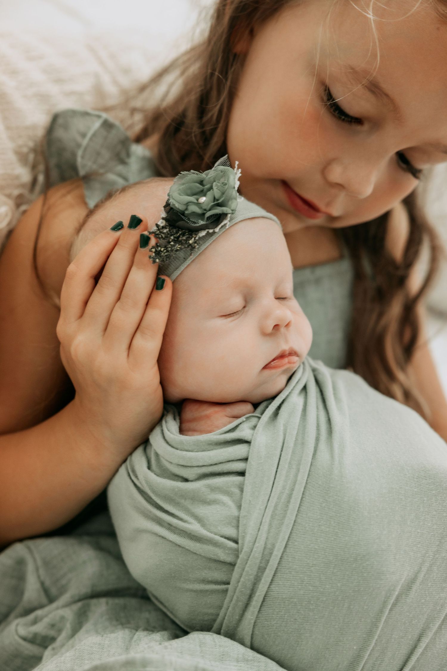 Older sister gently holding sleeping newborn baby wrapped in sage green swaddle with floral headband.