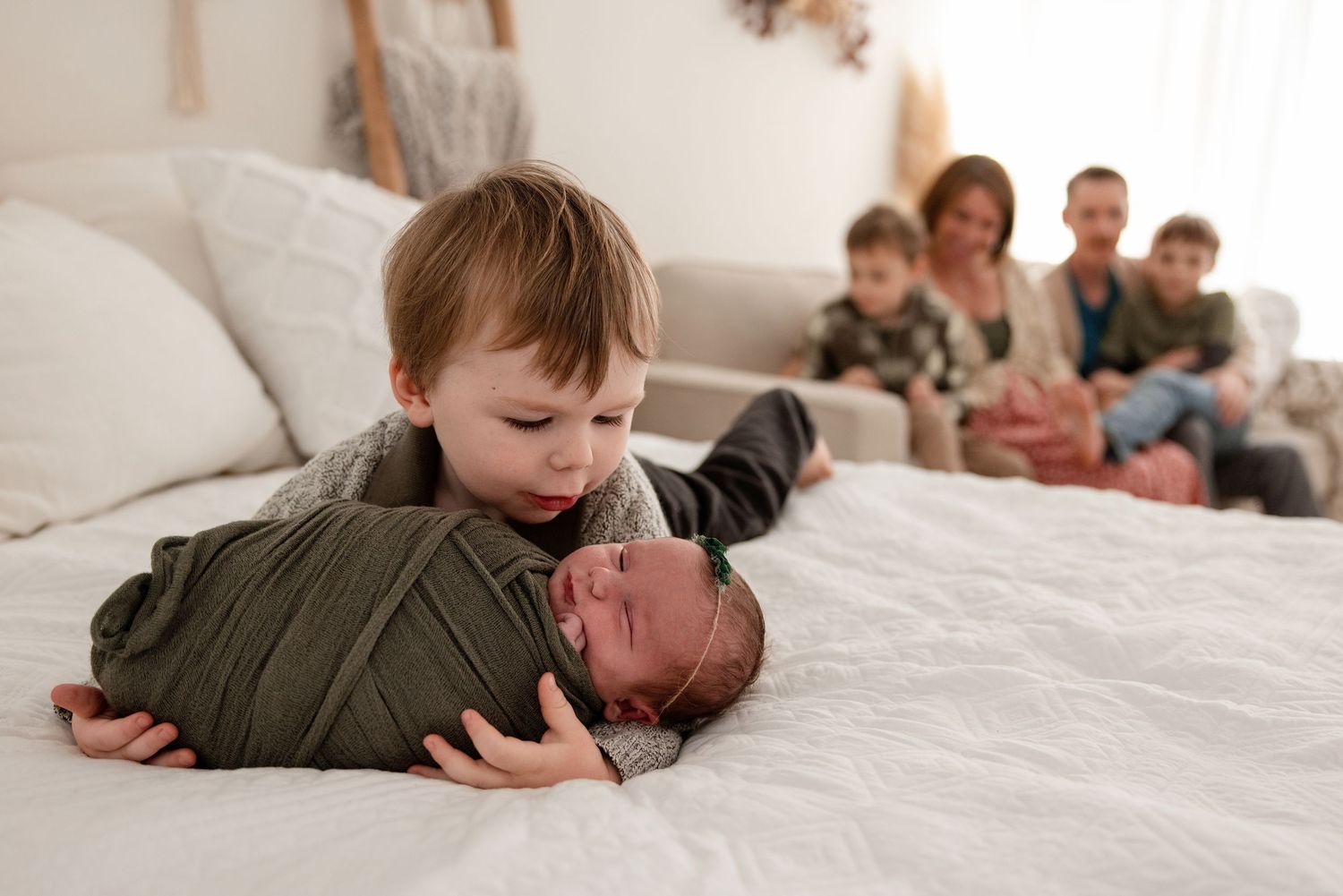 A toddler boy gently holds a swaddled newborn on a white bed while family members watch warmly in the background.