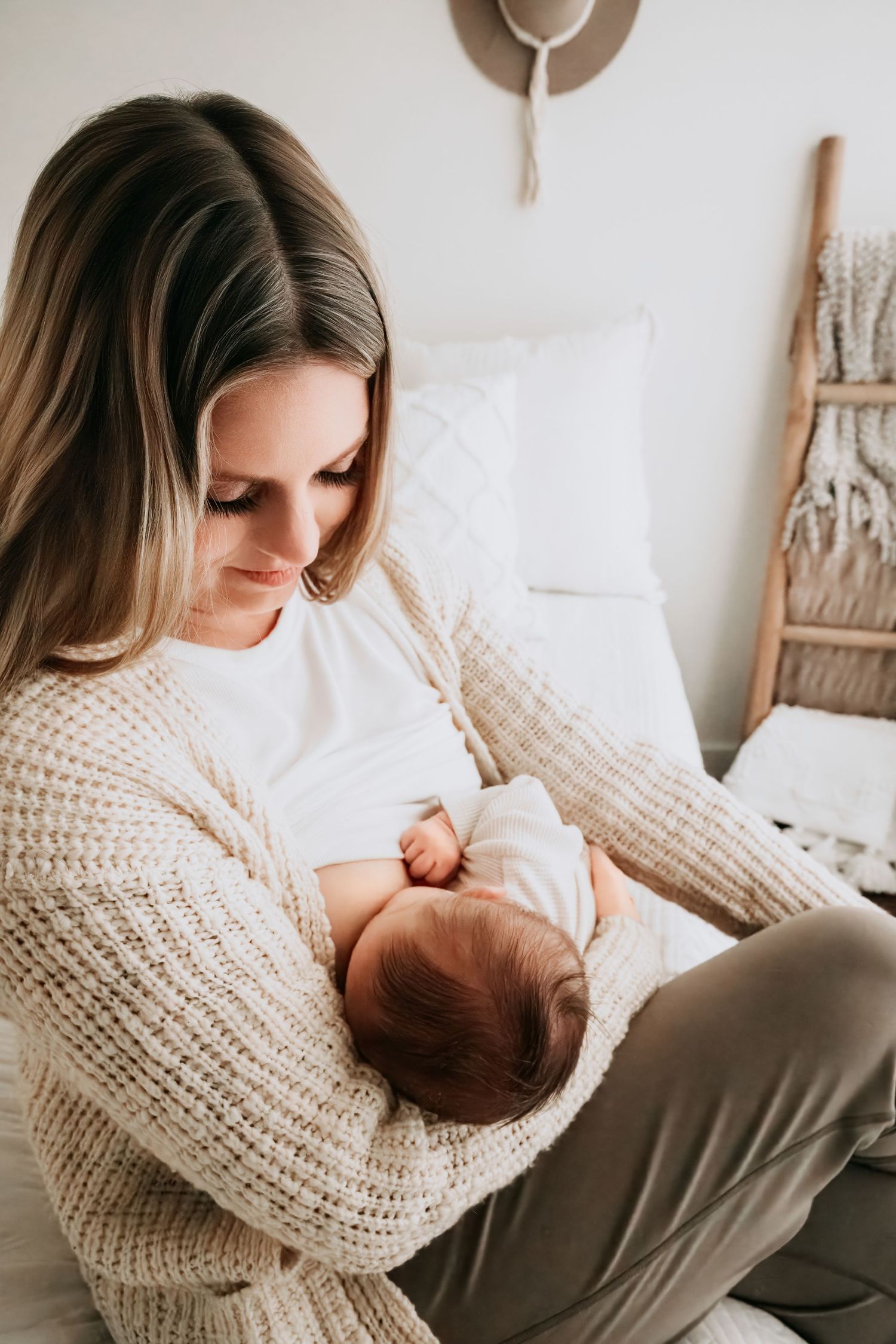 A young mother in a cozy knit cardigan tenderly breastfeeds her newborn baby while sitting in a softly lit bedroom.