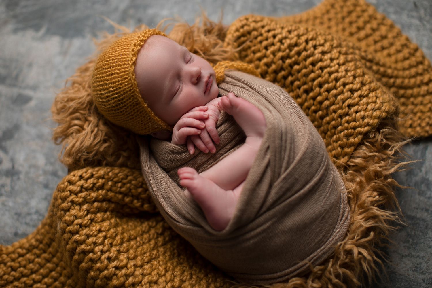 Newborn baby swaddled in tan wrap with mustard knit bonnet on golden textured blanket.  at The Studio Bonsall, near Vista and Oceanside