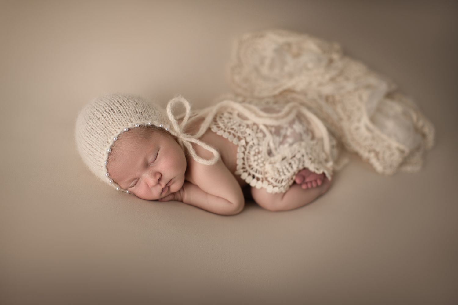 Sleeping newborn baby wearing cream knit bonnet posed with lace wrap on beige backdrop.