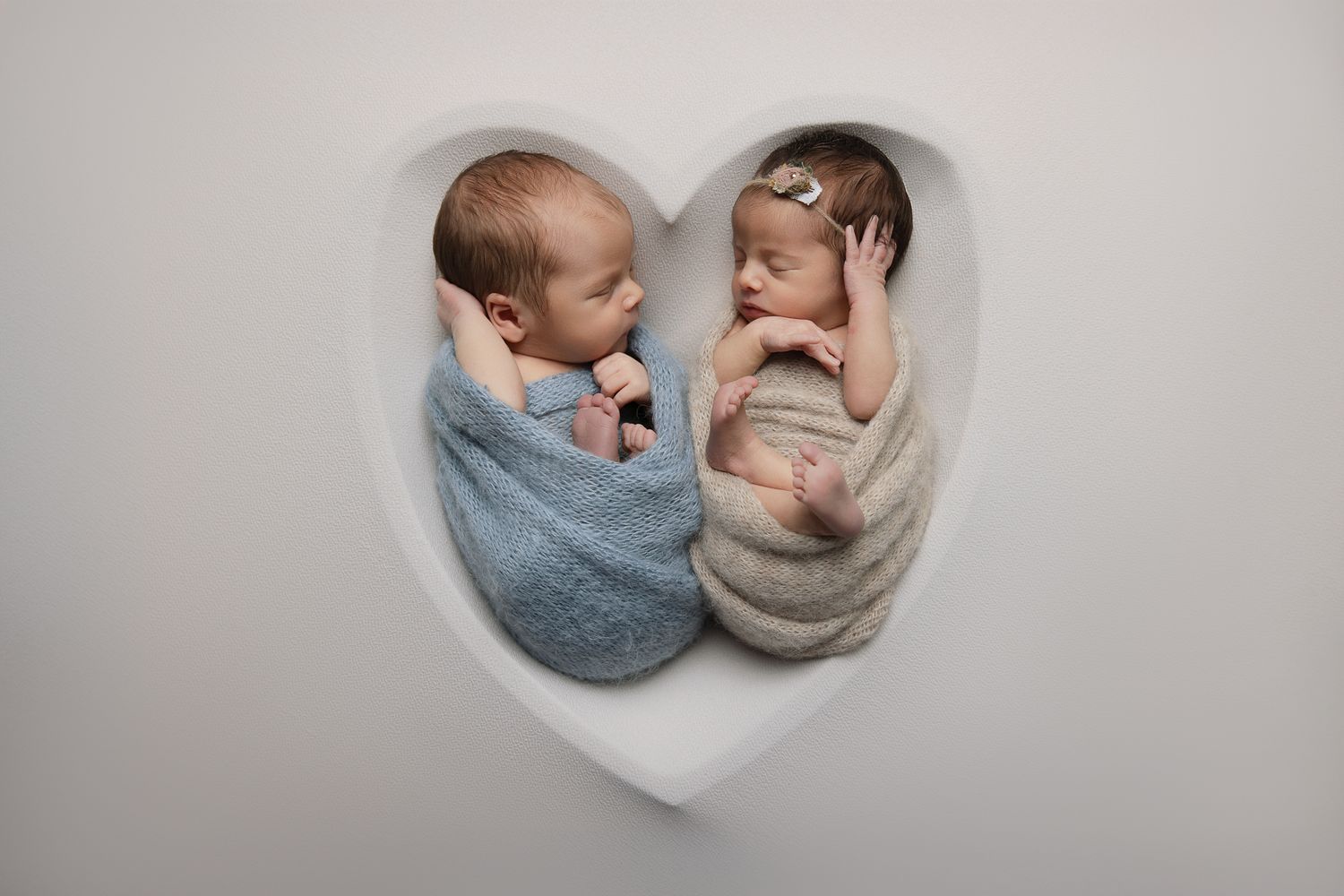 Two sleeping newborn twins swaddled in blue and beige wraps nestled together in a white heart-shaped prop.