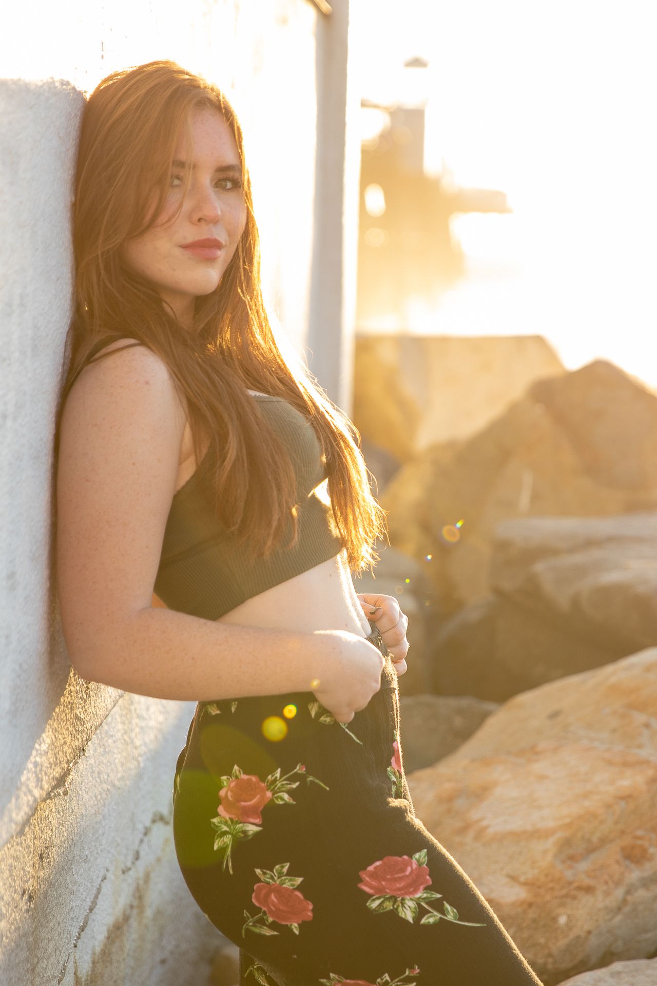 Pregnant woman with long red hair poses against a wall at sunset near rocky beach in floral skirt. Senior photography session in Oceanside