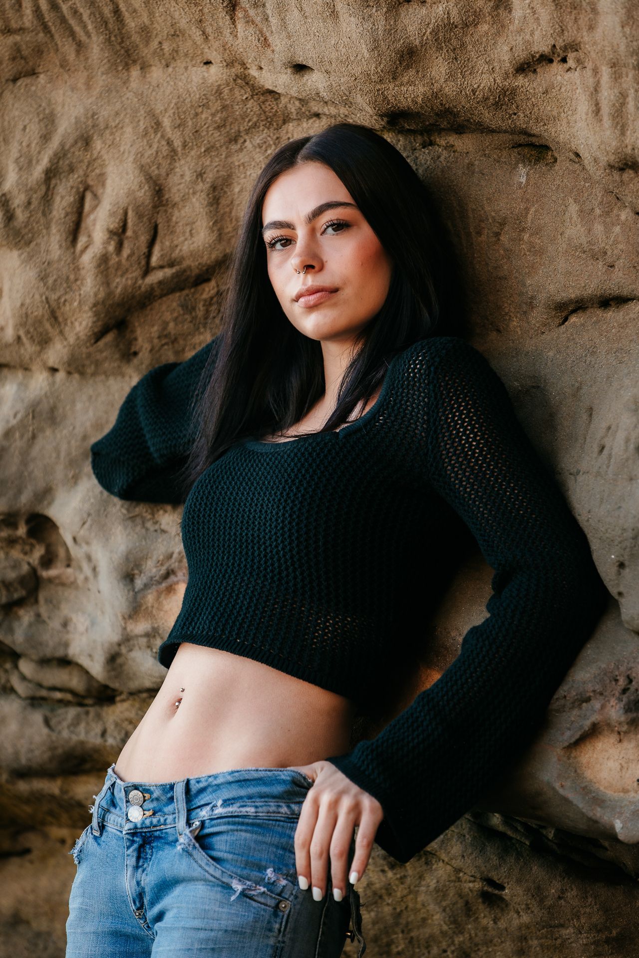 Dark-haired woman in black crop sweater and jeans leans against rocky stone wall.