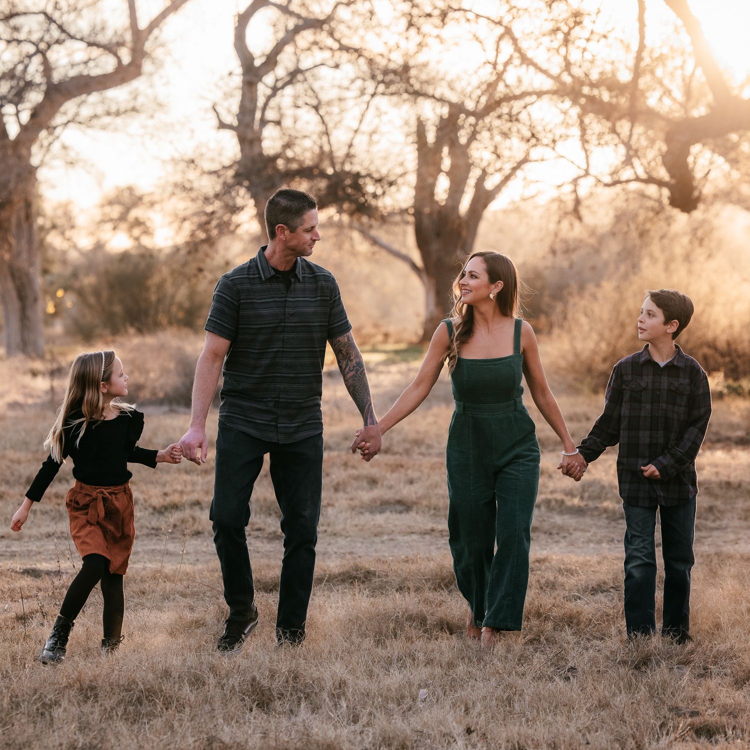 Family of four holding hands walking through golden field at sunset with autumn trees at the Bonsall Preserve.