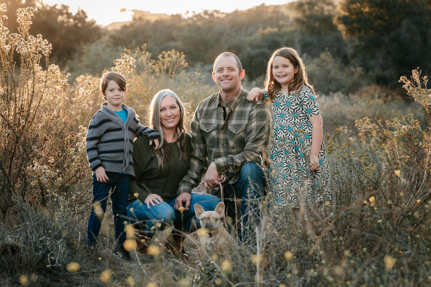 A family of four smiles together in a sunlit field of wildflowers during a warm autumn afternoon in Bonsall CA