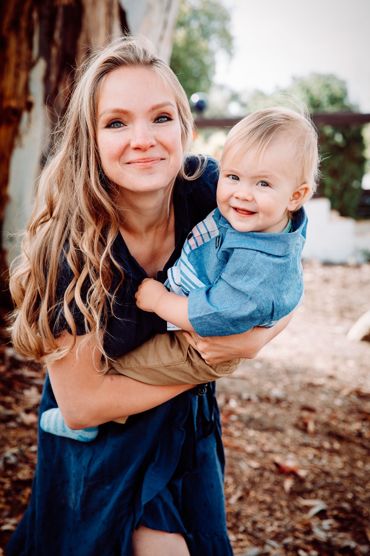 Smiling blonde woman holding an adorable baby boy in a blue shirt during an outdoor portrait session.