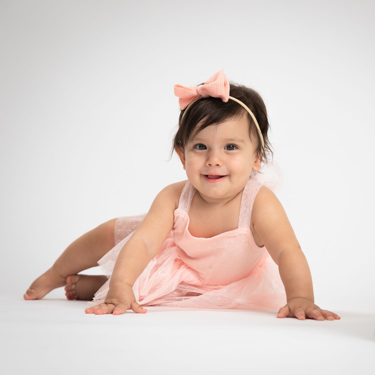 Smiling baby girl in pink dress and bow headband posing on white studio floor.