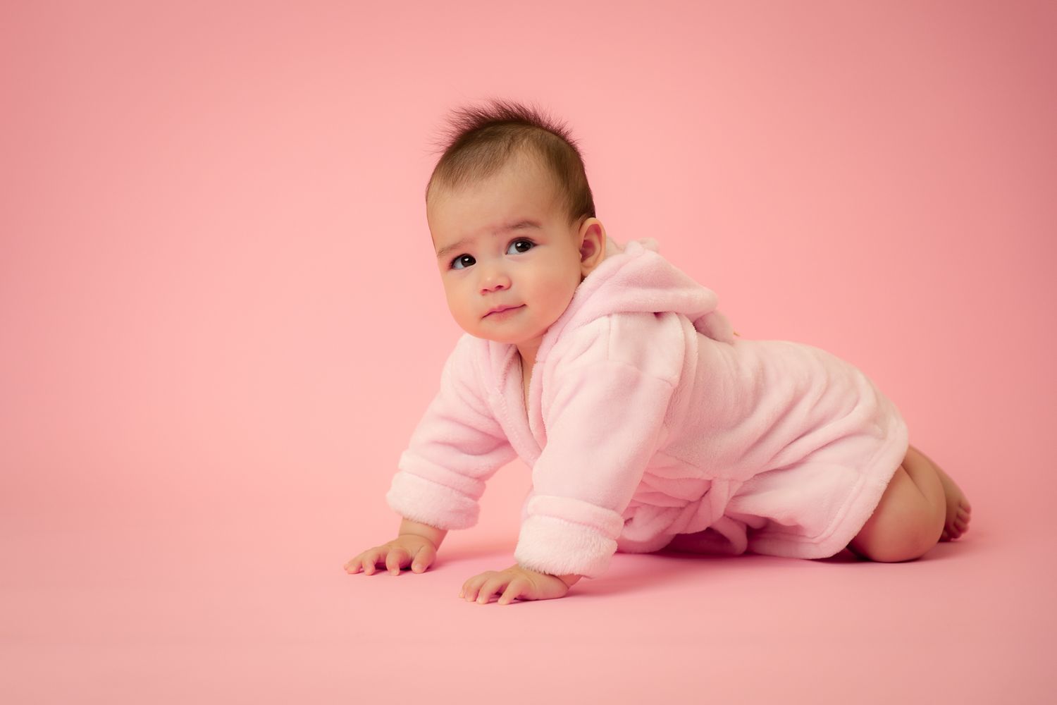 Cute baby in pink fluffy robe crawling on a pink background.