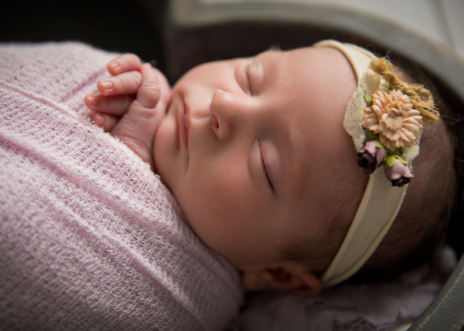 Peaceful newborn baby girl sleeping swaddled in pink wrap wearing a floral headband.
