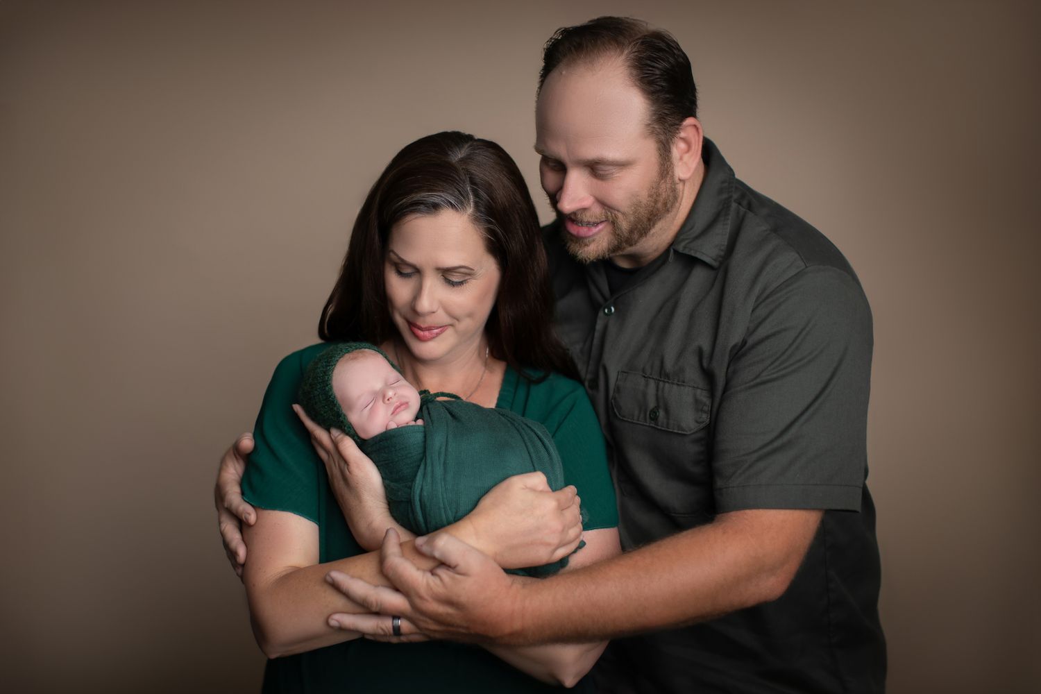 A couple in green and gray outfits tenderly holds their sleeping newborn baby during a professional studio portrait session.