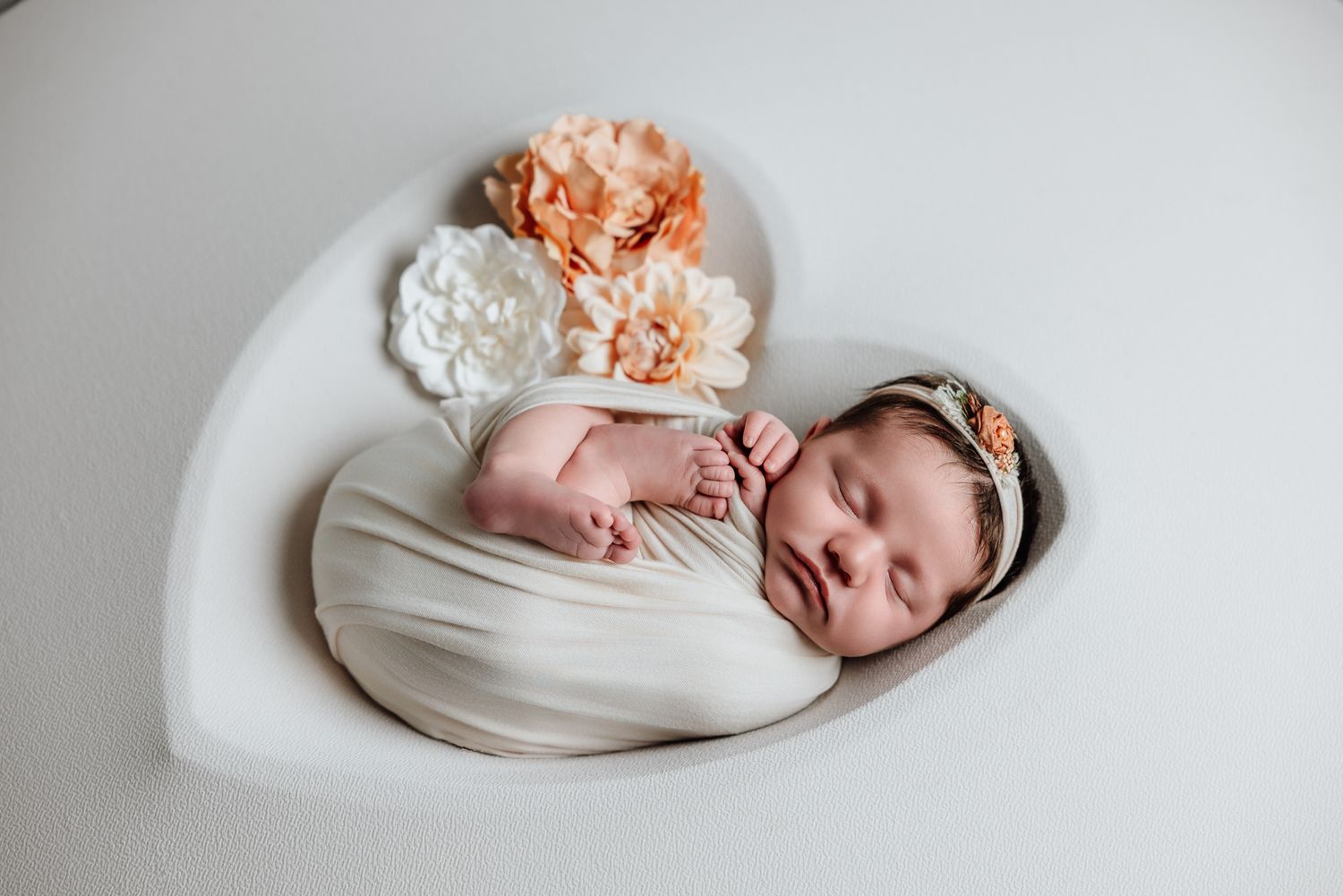 Sleeping newborn baby swaddled in cream wrap inside heart-shaped prop with peach and white flowers.