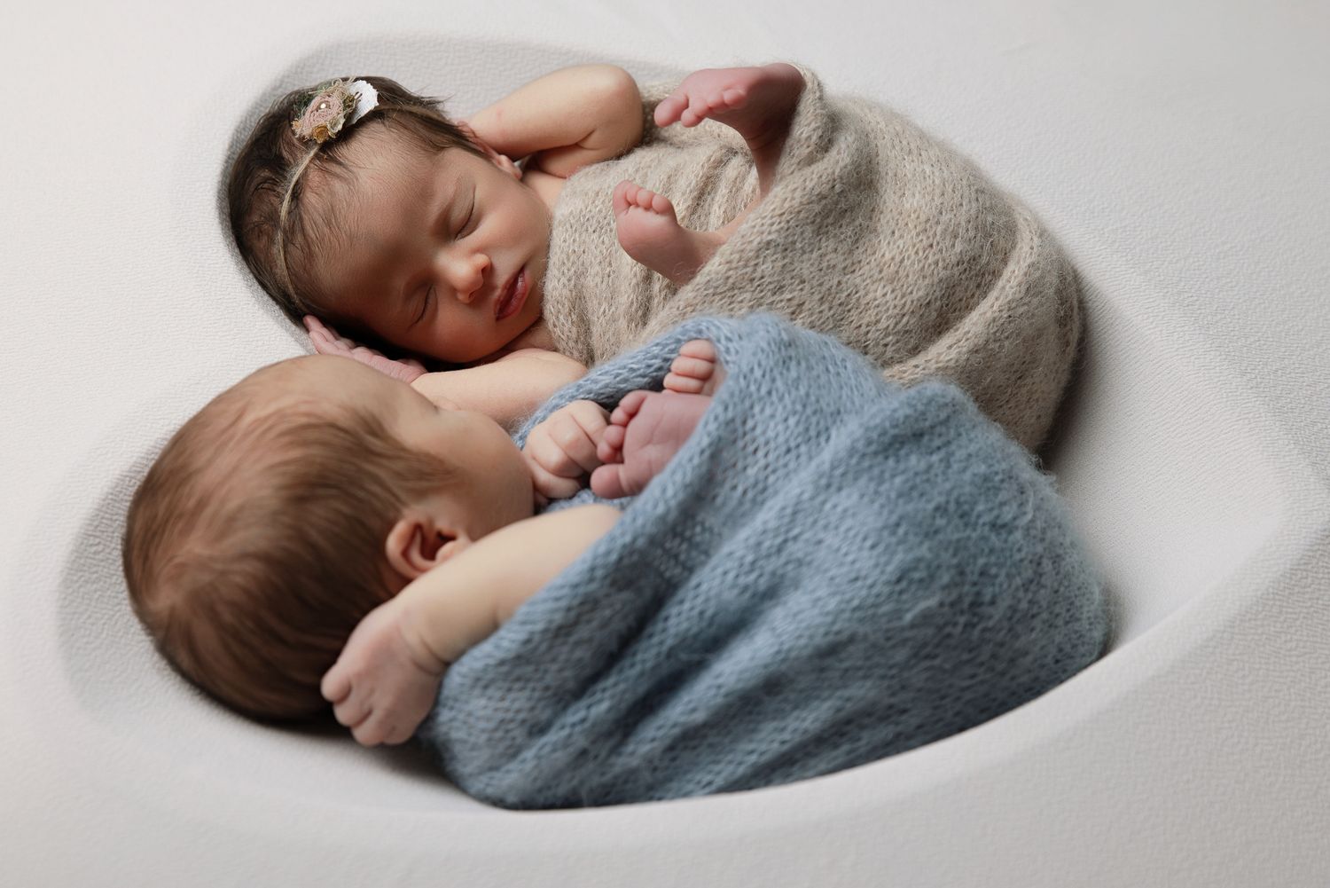 Two sleeping newborn twins swaddled in gray and blue wraps curled together in white bowl.
