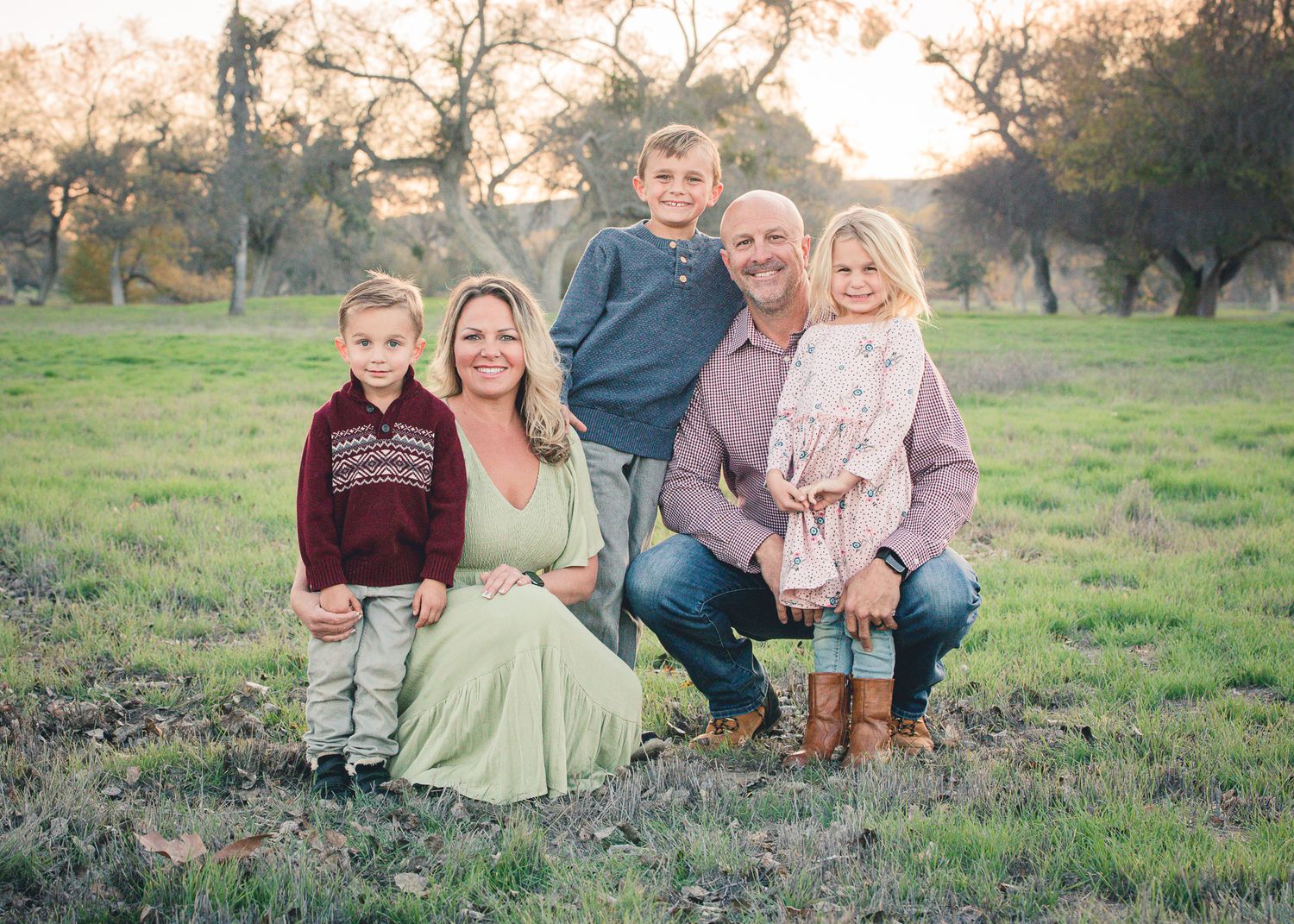 Family of five posing in an open green field at golden hour with warm sunset light behind them.
