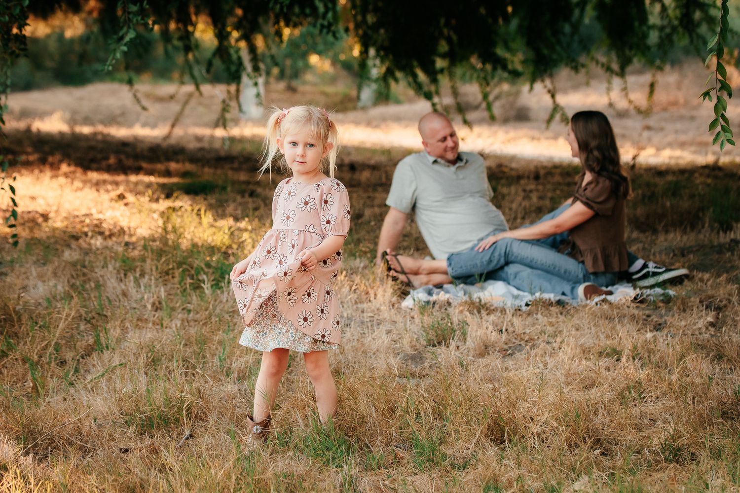 Young blonde girl in pink dress standing in field while parents relax on blanket behind her.
