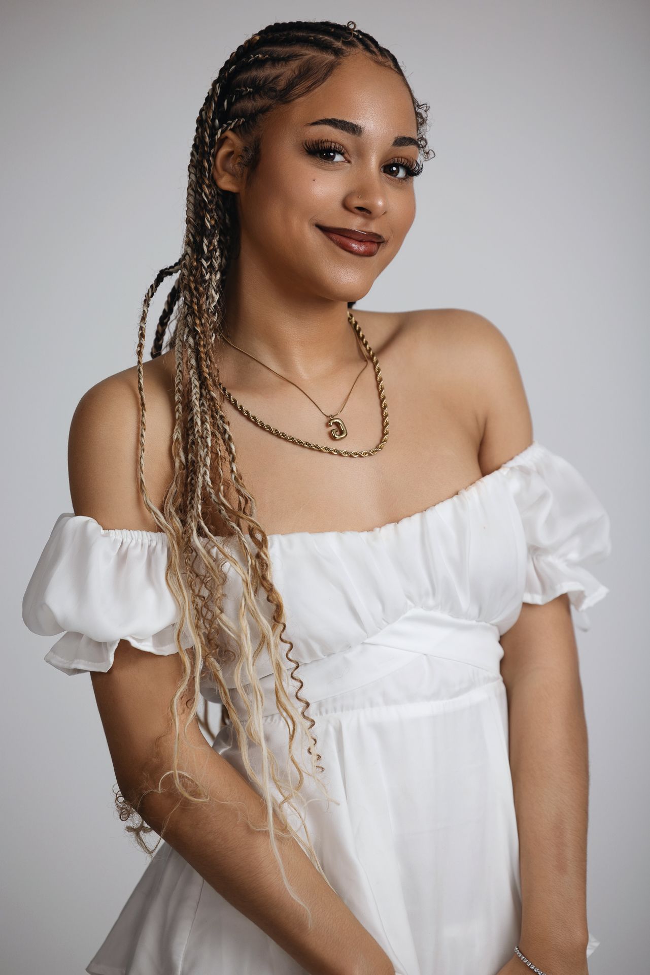 Smiling young woman with long braids wearing white off-shoulder dress and gold necklaces in studio portrait.