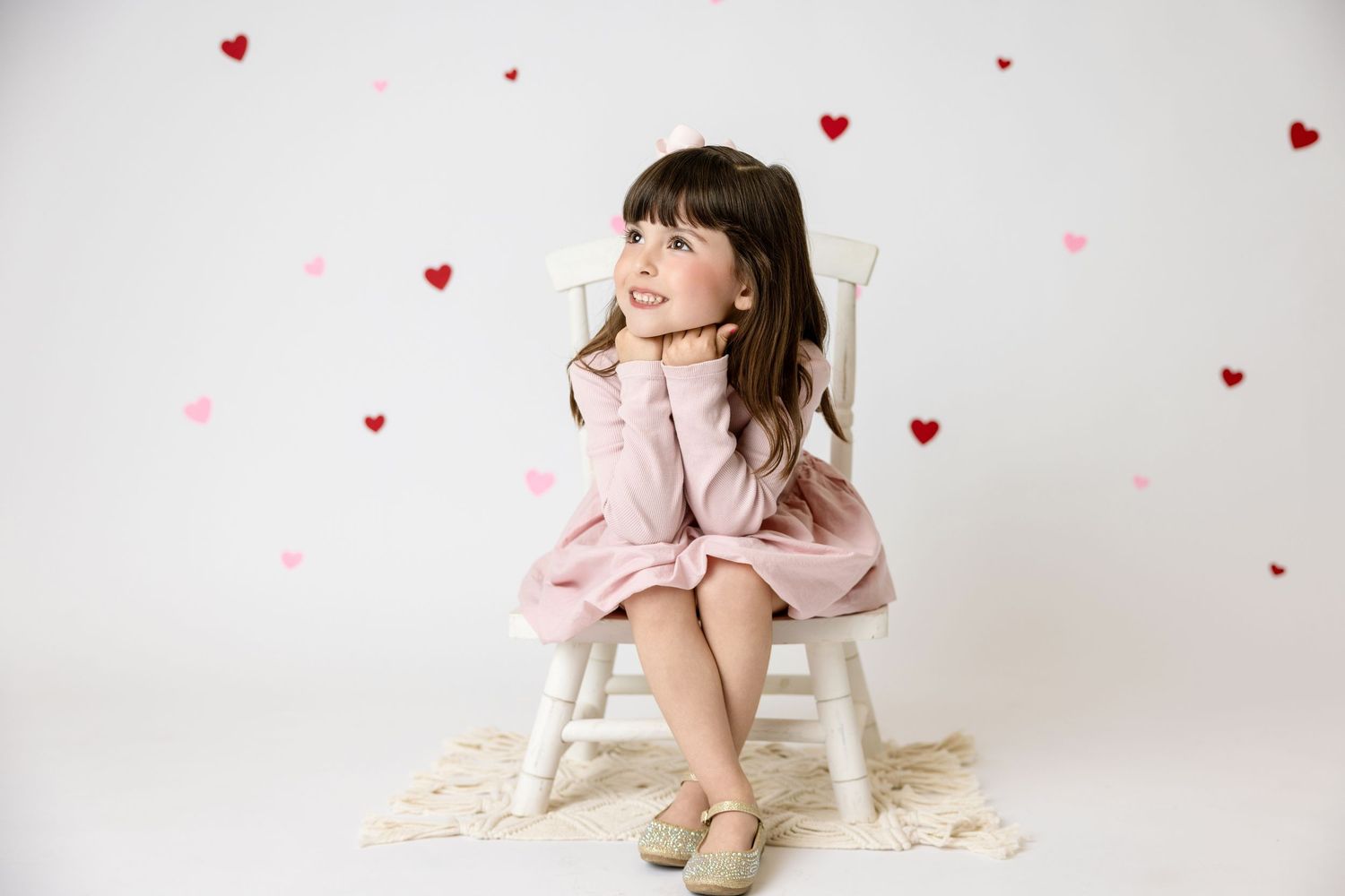 Adorable young girl in pink dress sitting on white chair surrounded by red and pink heart decorations. in the Bonsall Studio near Fallbrook,