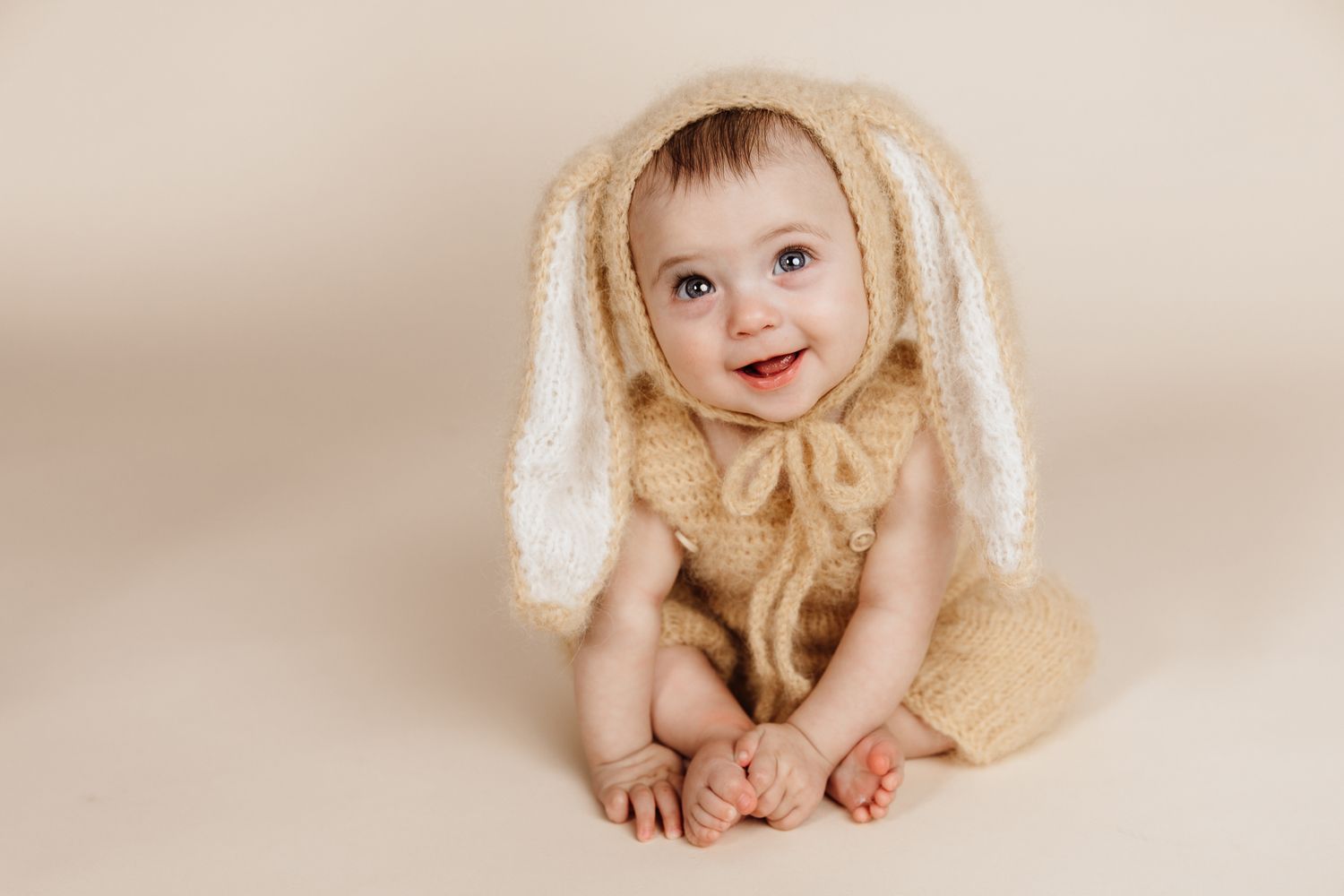 Smiling baby wearing a knitted bunny ear costume in cream color, sitting on a beige background.