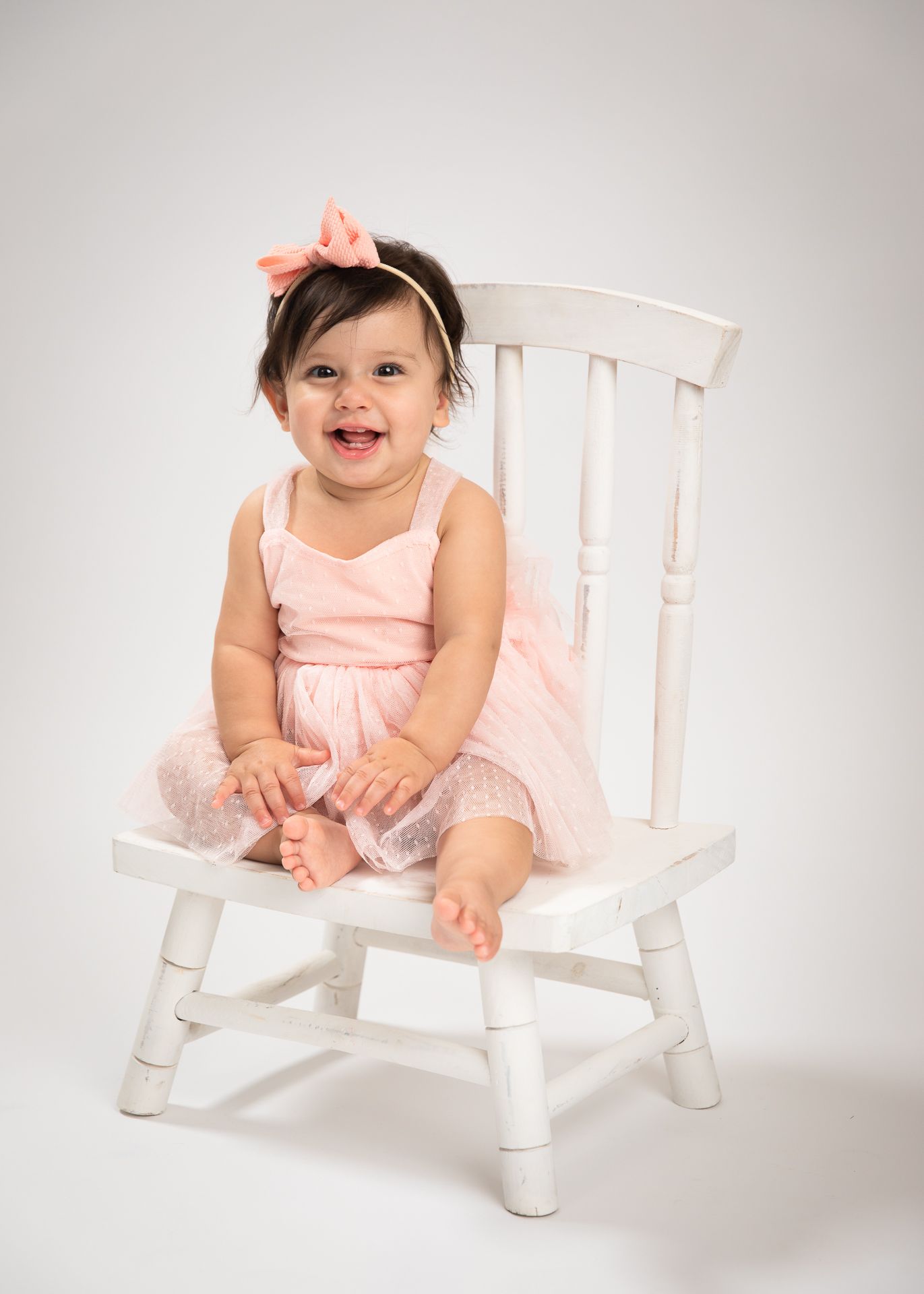 Happy baby girl in pink tutu dress sitting on white wooden chair with bow headband.