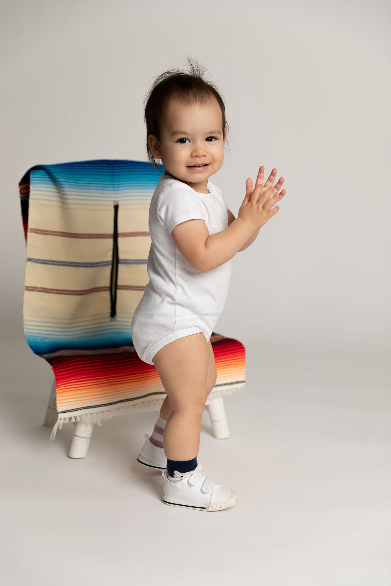 Happy baby in white onesie waving while standing by a colorful striped chair.
