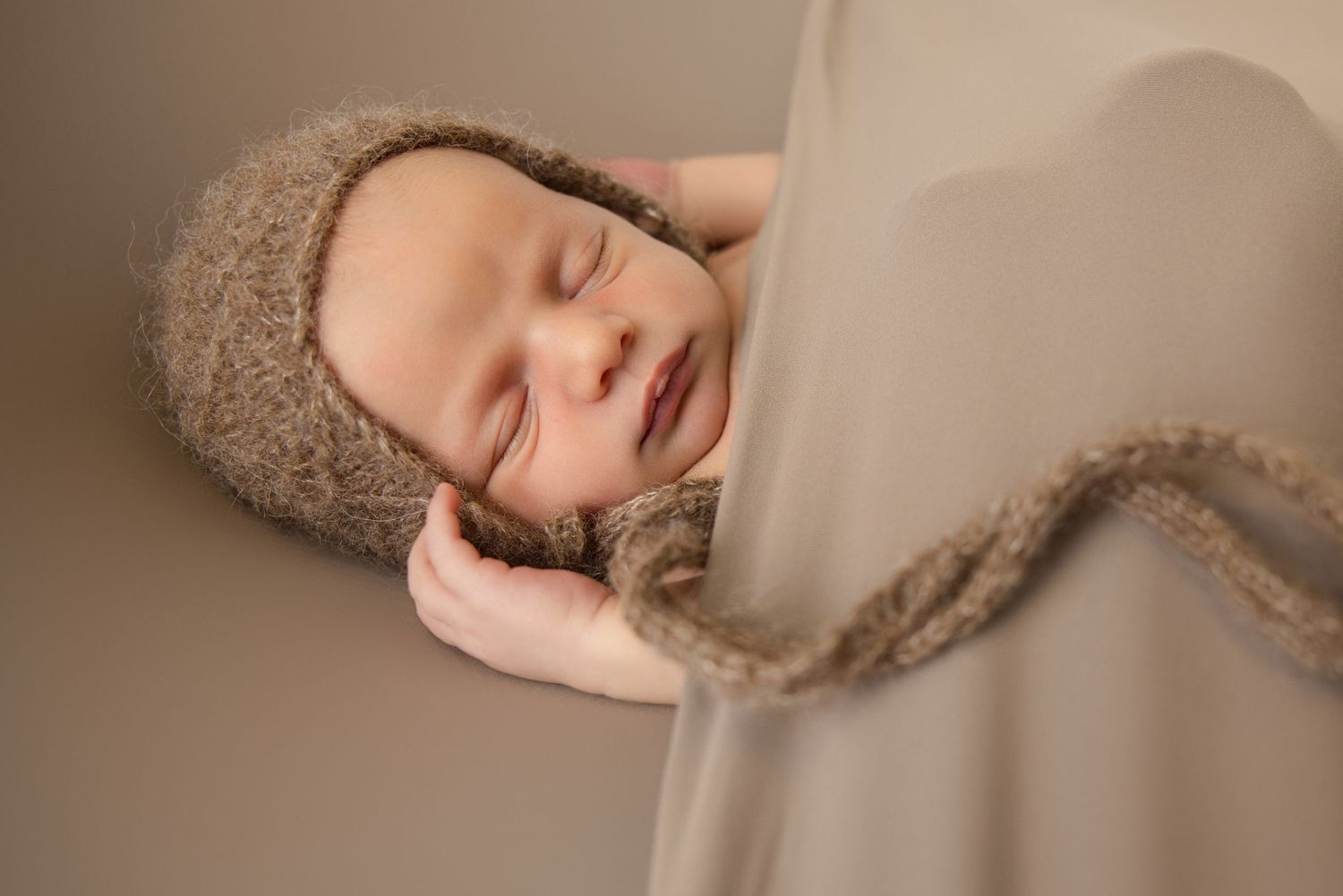 Sleeping newborn baby wearing a cozy brown knit bonnet, peacefully resting on a soft neutral backdrop.