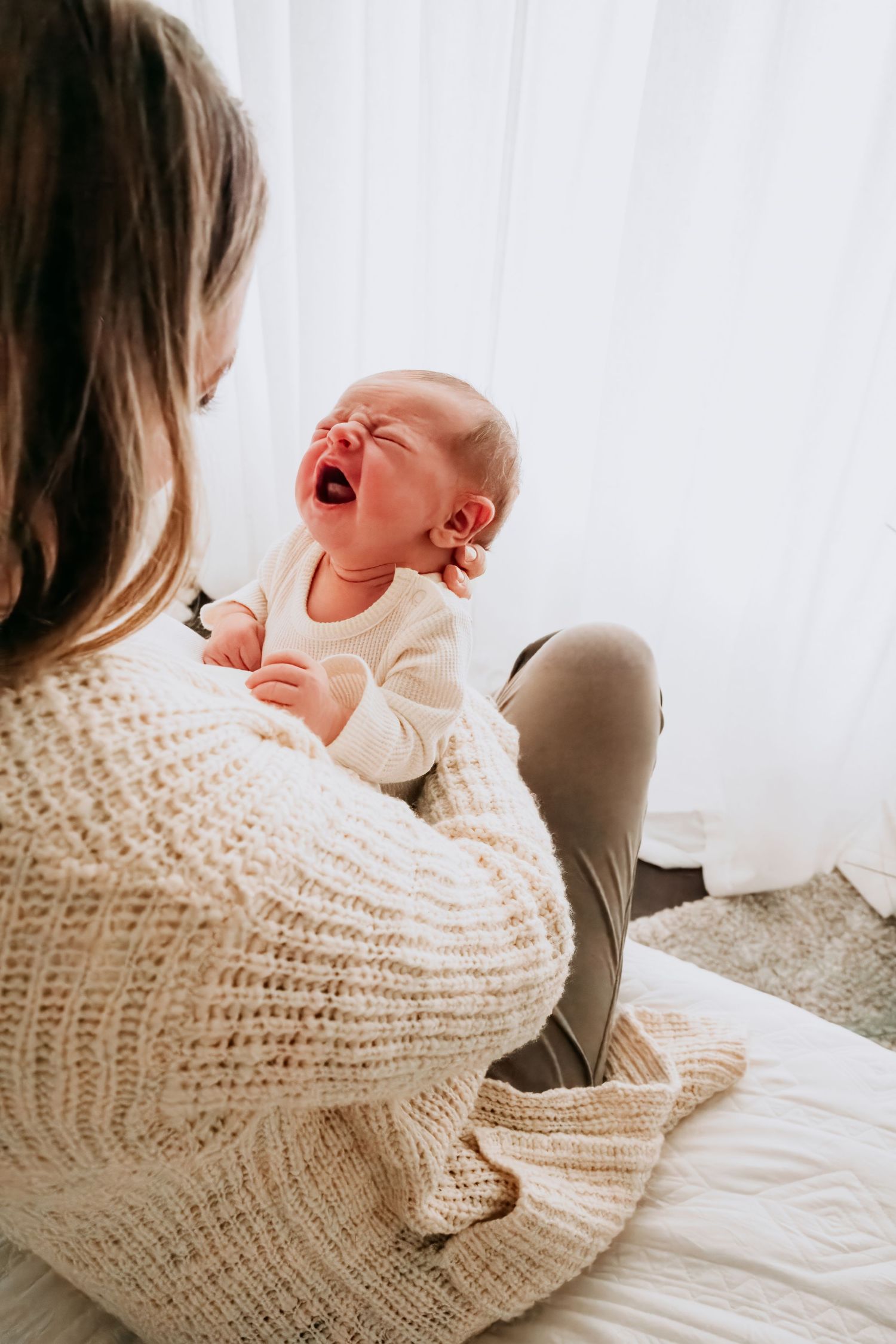 A mother in a cozy knit sweater holds her crying newborn baby while sitting on a bed near a bright window.