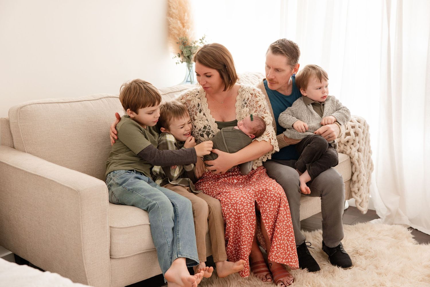 Family of five sitting on a cozy beige sofa, parents with three young children including a newborn baby.