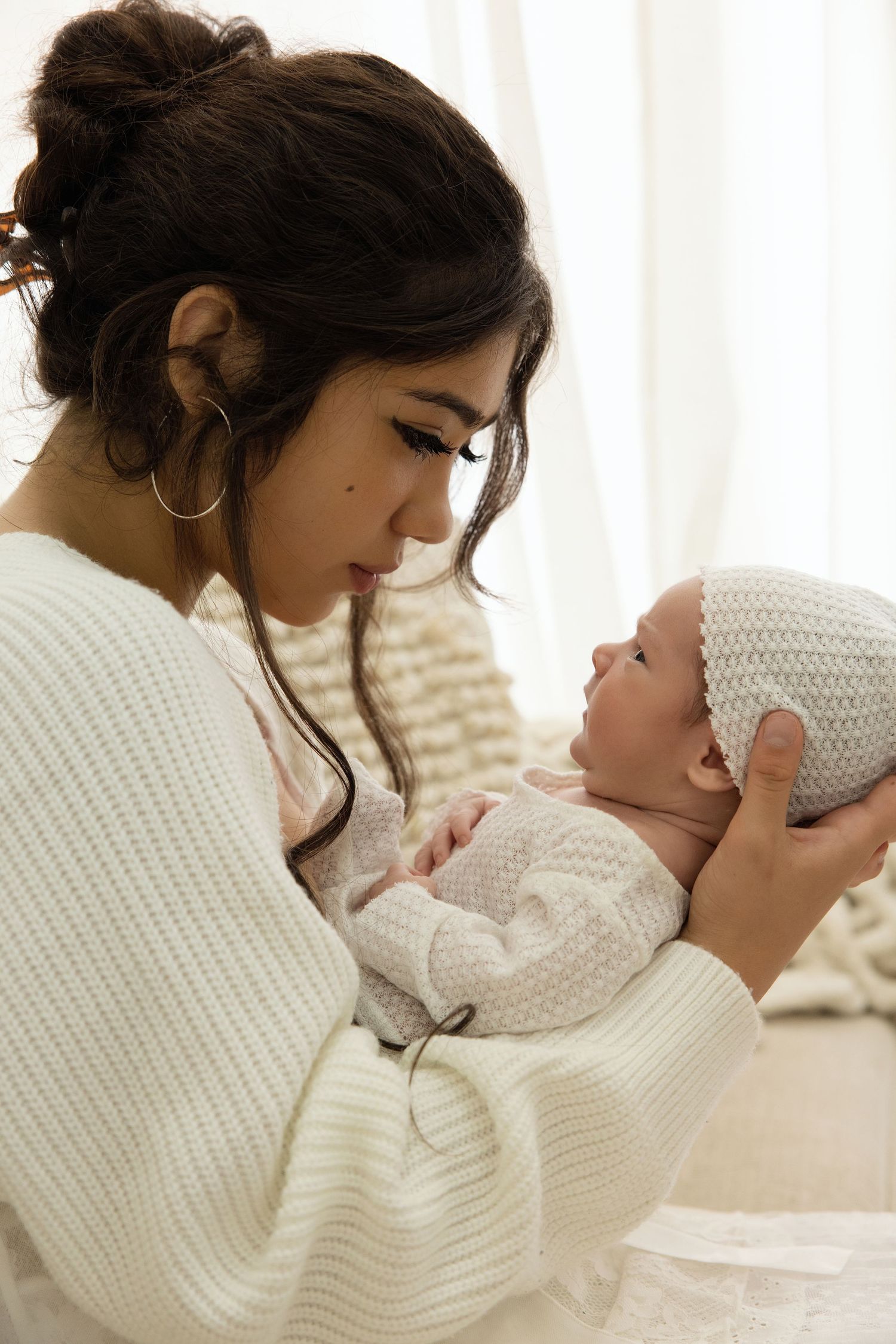 A young mother in a white sweater tenderly holds her newborn baby wearing a knit hat near a bright window.