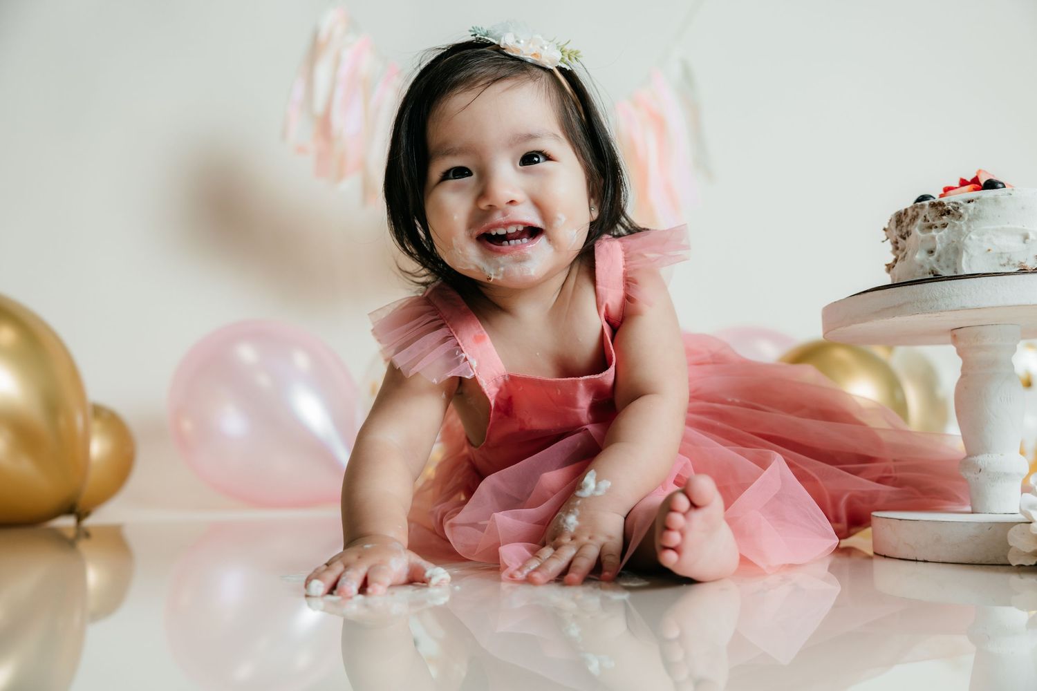 Happy baby girl in pink tutu dress crawling near birthday cake and colorful balloons during cake smash photoshoot.