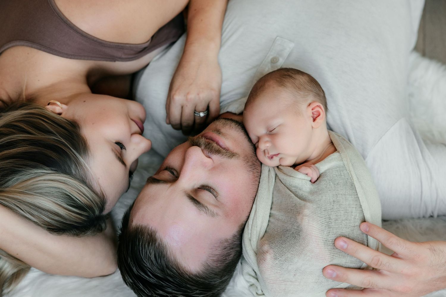 New parents lying down with eyes closed, lovingly cradling their sleeping newborn baby wrapped in a soft blanket.