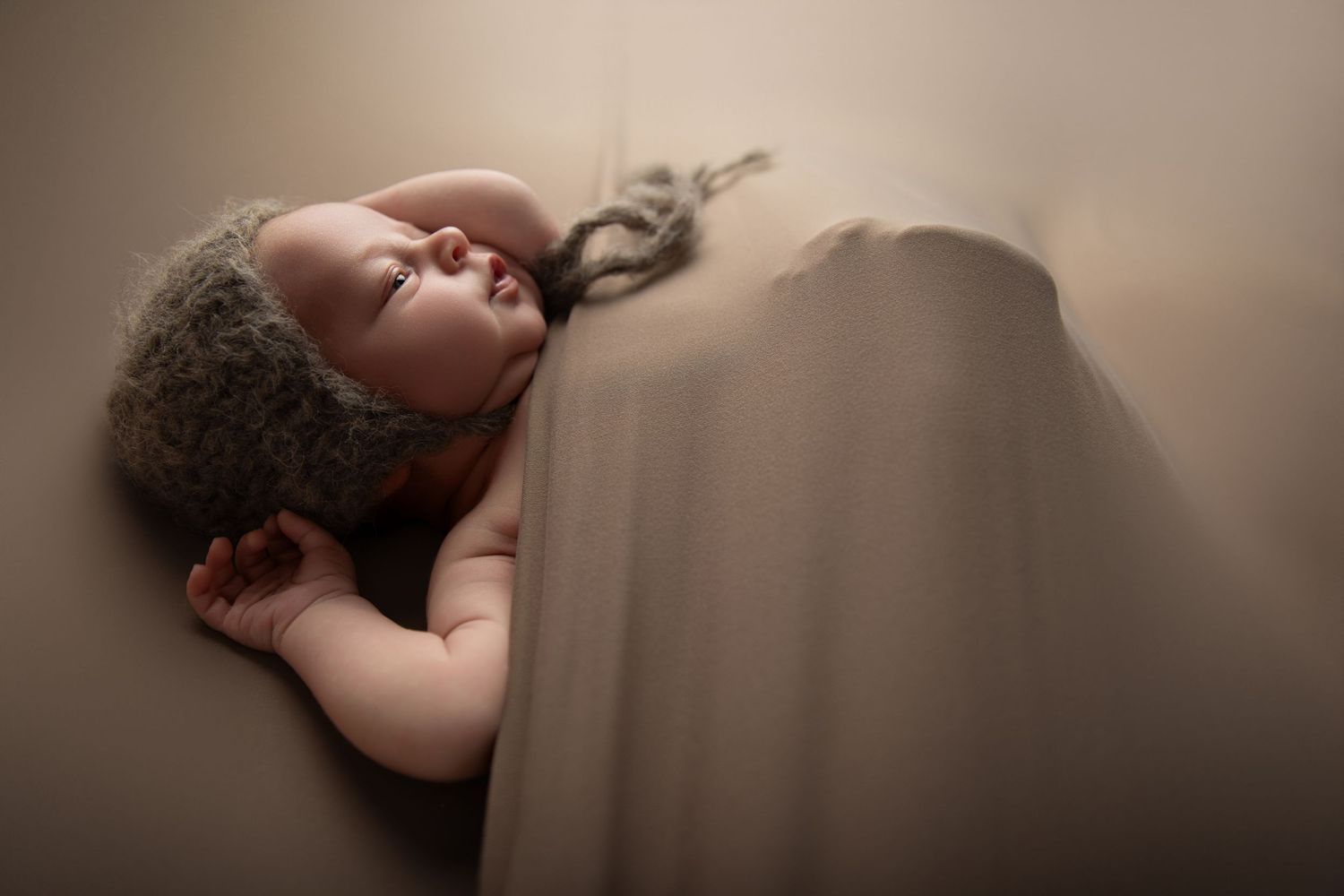 Newborn baby sleeping peacefully wrapped in a soft beige blanket wearing a cozy knit hat in a studio portrait.